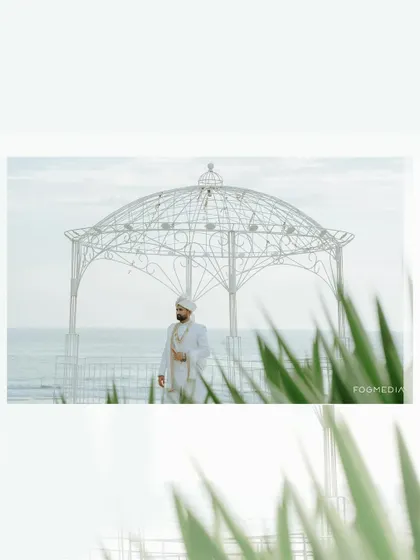 The groom stands by a white gazebo on the beach, a handsome and serene portrait before his wedding.