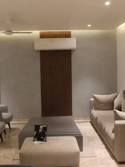 This view of the Hyderabad living room showcases the clean lines and balanced layout. A textured gray wall is complemented by a dark wood panel, creating a subtle feature behind the seating area. Cove lighting provides a soft, ambient glow throughout the space.