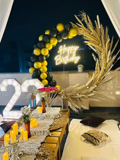 A close-up of a stylish birthday dinner arrangement on a terrace. This setup features a 'Happy Birthday' neon sign, a modern balloon arch with pampas grass, and a beautifully set low table with a macrame runner and candles.
