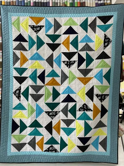 A "Twisted Geese" quilt with a playful, modern aesthetic. The random arrangement of flying geese blocks in cool tones of blue, green, and yellow creates a sense of movement.