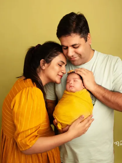 A new family of three, wrapped in love. The parents, dressed in warm yellow tones that match the baby's swaddle, gently hold their newborn in this tender studio portrait.