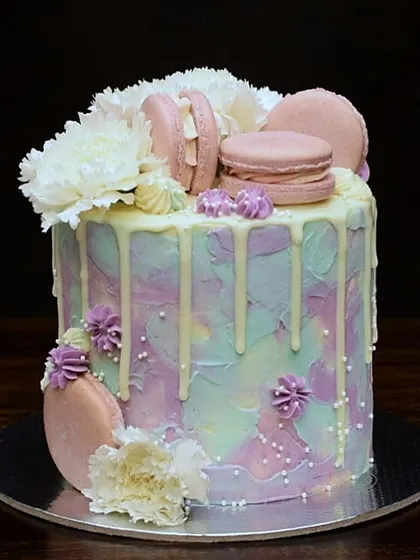 A beautiful cake with a pastel watercolor buttercream finish. It's adorned with fresh white carnations, pink macarons, and a white chocolate drip for a soft and dreamy look.
