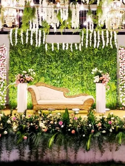 This reception stage combines natural and elegant elements, with a dense greenery wall as the centerpiece. The design is accented with panels of white and pink flowers and a ceiling of hanging white florals, creating a garden-like feel indoors.