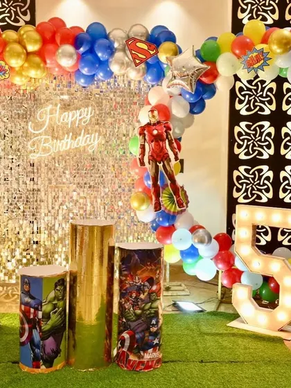 An action-packed Avengers-themed 5th birthday party. The setup features a silver shimmer wall, a colorful balloon arch, character props, and a light-up number '5'.