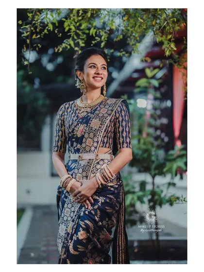 A smiling portrait of Prathima. Her makeup is subtle and sophisticated, perfectly complementing her intricately woven silk saree and traditional jewelry.