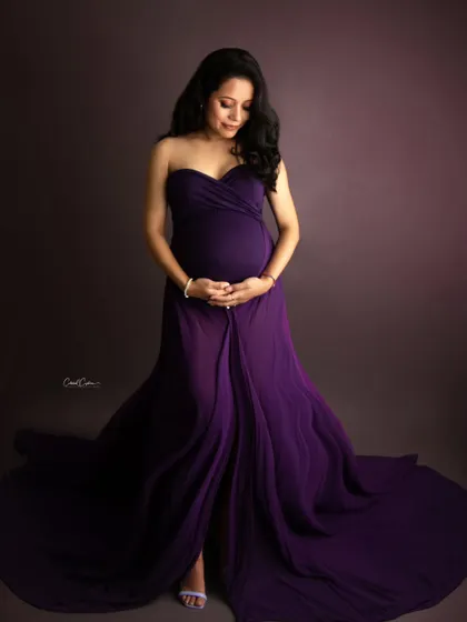 A classic and elegant studio portrait. The mom-to-be looks down, cradling her bump in a simple yet stunning deep purple gown.