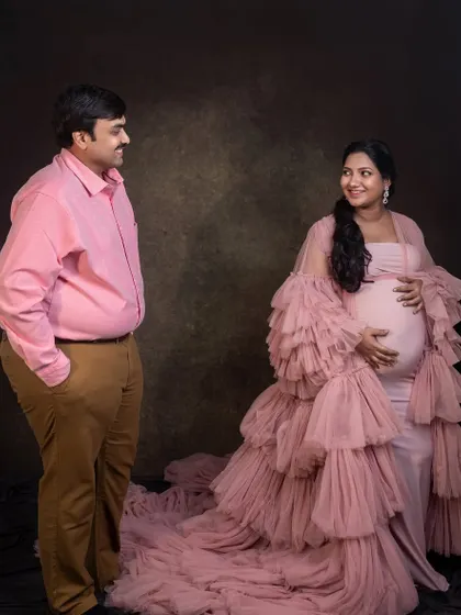 An expecting couple looks at each other with love during their studio maternity session. The mother wears a dramatic pink ruffled gown, adding a touch of glamour and style to the portrait.
