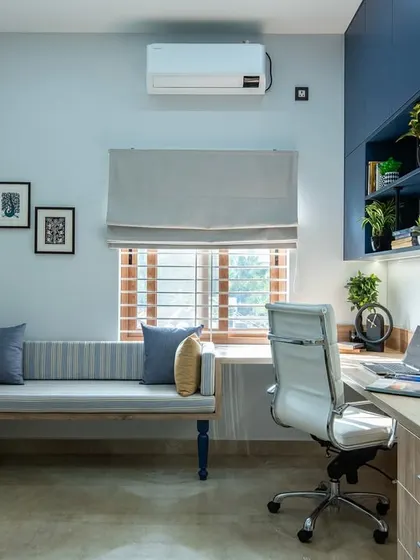 A clean and organized study nook with a wooden desk, a white ergonomic chair, and a simple grey bench. The design is minimalist yet functional, with framed art and Roman blinds adding a touch of personality.