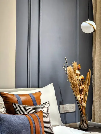 A close-up of a bedroom corner, showing a mix of textures and colors. Leather and fabric cushions with geometric patterns sit against a deep blue paneled wall. A vase of dried flowers on the nightstand adds a natural, rustic touch.