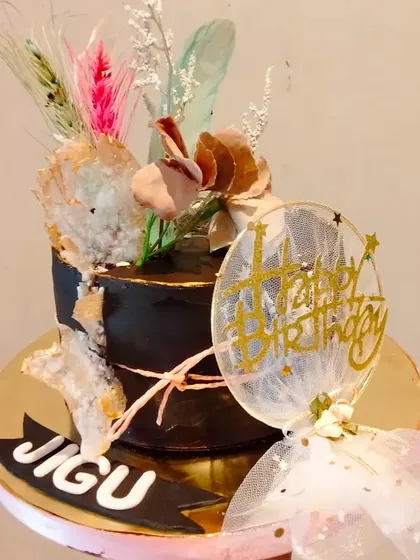 A chic black cake with bohemian-style decorations. The design features rice paper sails and dried flowers, creating a modern and artistic look for a birthday celebration.
