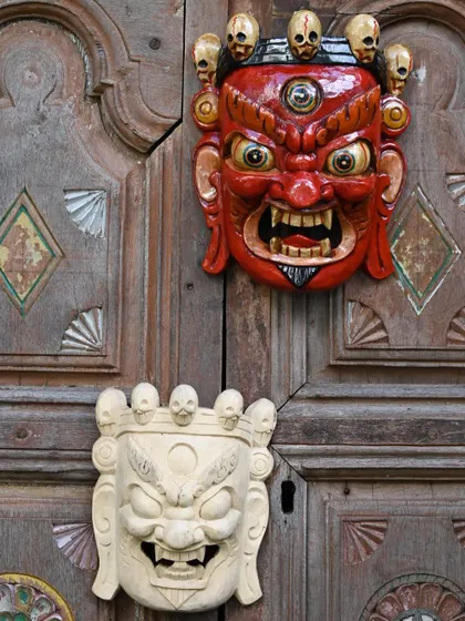 Two traditional masks from Nepal, one painted and one in a natural finish, displayed on a vintage wooden door. This shows the versatility of the art form, where each mask has a distinct personality.