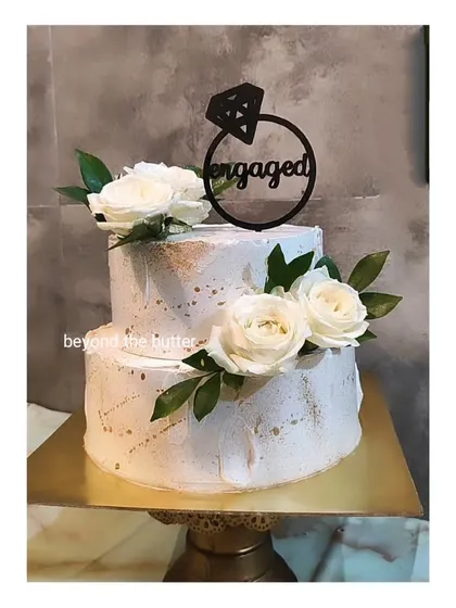 A modern and minimalist two-tier engagement cake. The textured white buttercream is splattered with gold, adorned with simple white roses, and finished with a bold black "Engaged" ring topper.