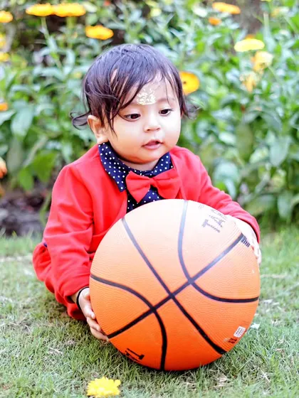 A young boy in a red suit jacket holds a basketball during a sports-themed outdoor photoshoot in a garden.