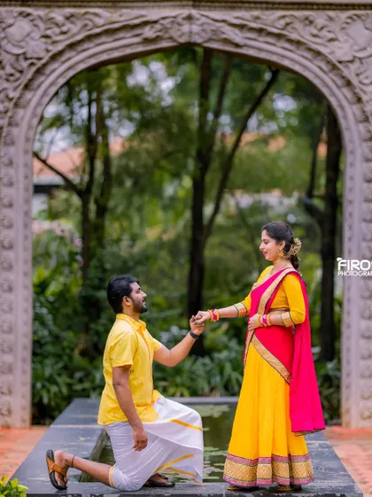 A playful and traditional proposal moment. The groom-to-be is on one knee, holding his partner's hand. Their colorful yellow and red traditional outfits look vibrant against the stone archway.