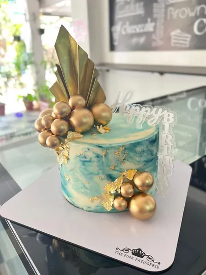 An artistic cake with a blue and white marbled effect, reminiscent of ocean waves. The design is finished with clusters of gold spheres, gold leaf, and a modern gold chocolate fan.