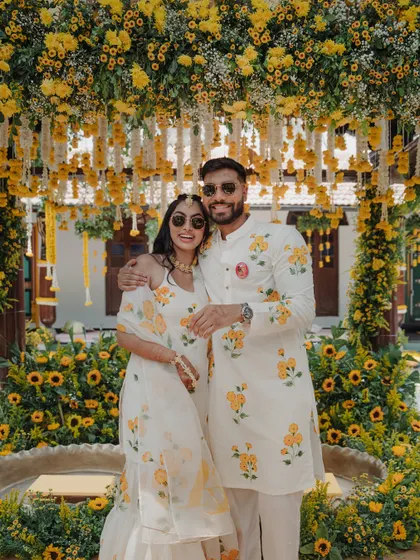 A happy couple poses in front of their stunning Haldi decor. Dressed in coordinated floral outfits, they are all smiles, surrounded by a sea of sunflowers and marigolds that perfectly match the joyous occasion.