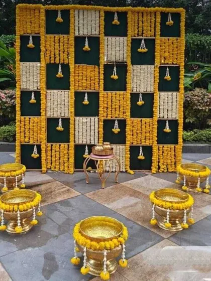 An elegant outdoor setup for a traditional ceremony. The backdrop is a grid of real marigold and tuberose flower strings, creating a beautiful photo opportunity. The space is accented with brass urlis filled with flowers.