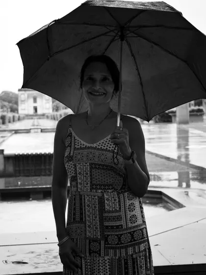 A black and white portrait of a woman smiling under an umbrella on a rainy day. The monochrome tones highlight her expression and the simple joy of the moment.
