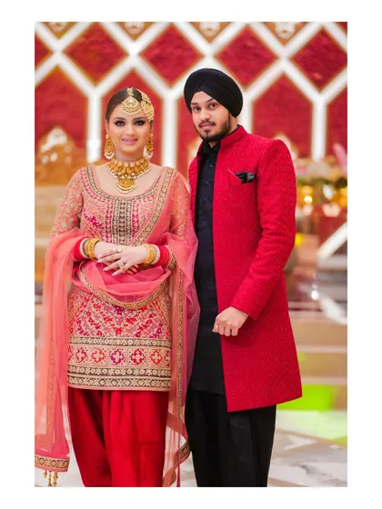 A classic portrait of a Sikh couple in their reception attire. The bride's red and pink salwar suit and the groom's contrasting red and black sherwani create a vibrant and traditional look.