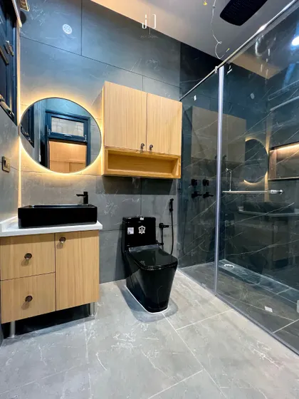 A modern bathroom design featuring a sleek black toilet and basin, complemented by light wood cabinetry. The backlit mirror and glass shower partition add a touch of hotel-like luxury and make the space feel open and bright.