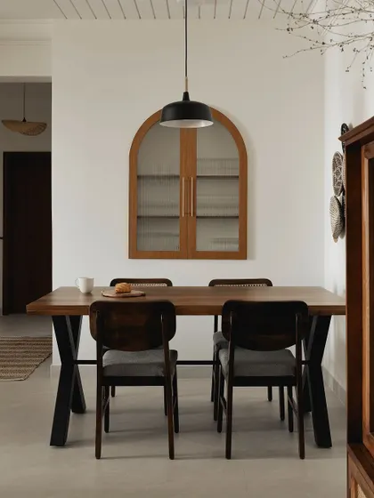 This dining room has a calm, Japandi-inspired feel. The dark wood table with its X-base is paired with simple chairs, and an arched, glass-front cabinet on the wall provides a beautiful display for crockery.