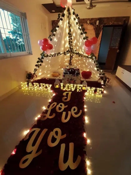 A full view of a romantic proposal or anniversary setup. A path of rose petals and lights leads to a cozy canopy tent, with 'I Love You' spelled out on the floor.