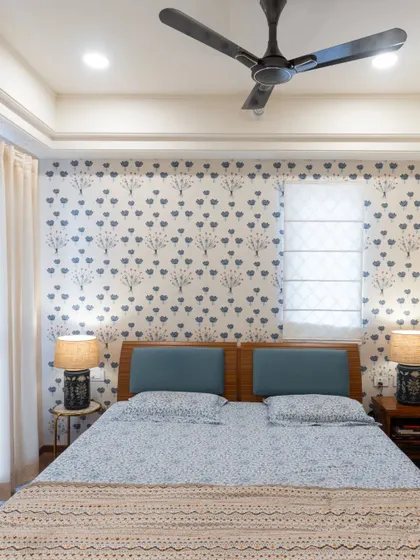 A full view of the bedroom with printed wallpaper and a simple wooden bed. The tray ceiling and neutral curtains add a touch of modern simplicity.