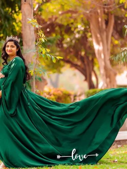 A majestic outdoor portrait of a mother-to-be in a flowing emerald green gown. The long train spreads out behind her, creating a stunning visual against the park's greenery.