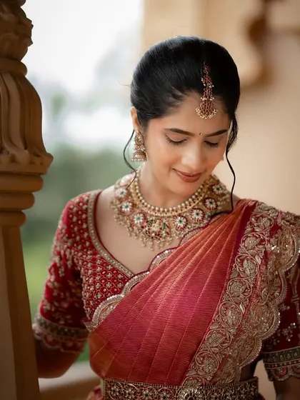A candid portrait of the bride, her gentle smile captured as she stands near a pillar in a grand venue. The soft light and her red saree create a warm and elegant image.