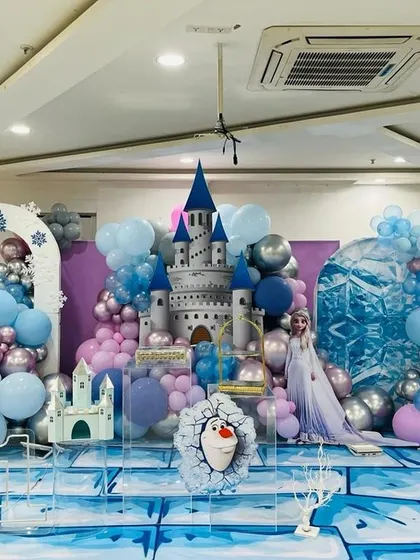 A grand Frozen themed party stage, featuring a 3D ice castle, character cutouts, and balloon arrangements that look like snow and ice.