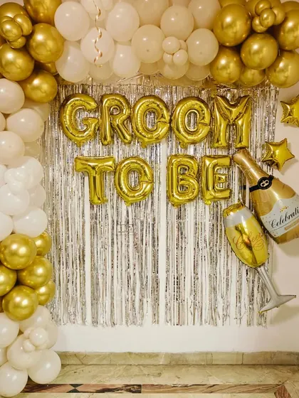A simple and stylish "Groom to Be" backdrop. This decoration uses a gold and white balloon arch against a silver fringe curtain.