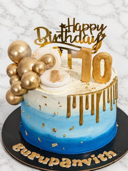 An elegant and modern birthday cake design. This cake features a blue ombre effect, a rich gold drip, and is decorated with golden chocolate balls and a touch of edible gold leaf for a sophisticated celebration.