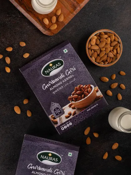 A top-down shot of Nauras Gurbandi Giri almond packaging on a dark, textured surface. The styling with bowls of almonds and milk suggests different ways to enjoy the product.