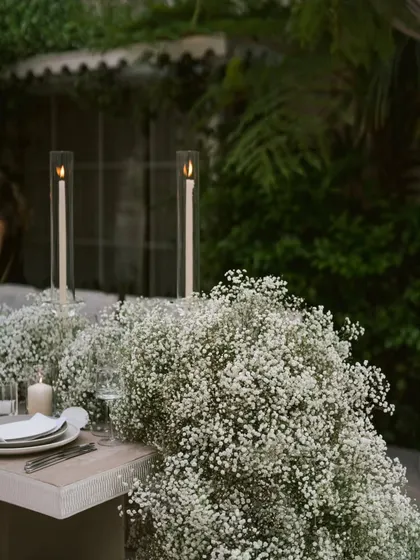 Tall, slender candles in glass hurricanes rise from a cloud of baby's breath on the dining table. This simple detail adds height and a touch of modern romance to the tablescape.