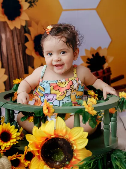 A happy baby girl sits in a green chair surrounded by sunflowers. This vibrant studio setup is perfect for a cheerful toddler photoshoot.