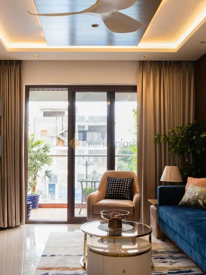 This living room combines a plush blue velvet sofa with a modern TV unit and a false ceiling with integrated cove lighting. The design creates a cozy yet sophisticated space with a great view of the balcony.