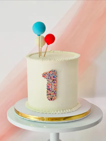 A simple and modern first birthday cake. The clean white buttercream is accented with a number "1" made of colorful sprinkles and three balloon toppers.