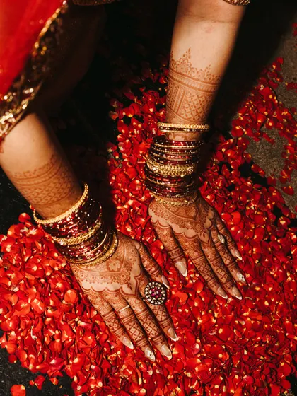 A close-up of the bride's hands, adorned with intricate mehendi and traditional red bangles, resting on a bed of rose petals.