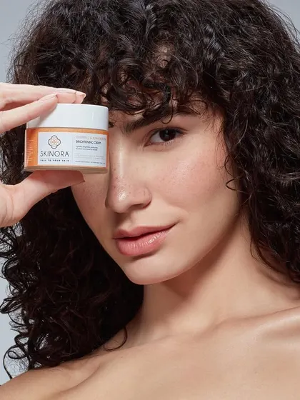 A product-forward portrait for Skinora Cosmetics. The model holds a small jar of face cream over one eye, drawing the viewer's attention directly to the product while maintaining a strong beauty aesthetic.