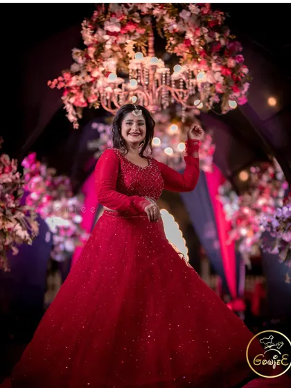The bride looking radiant in her red reception gown, posing under a floral chandelier.
