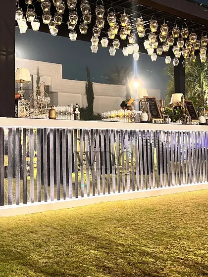 A sleek and modern bar setup. The bar front has a reflective, slatted design, and the overhead structure is adorned with dozens of hanging glass lanterns, creating a stylish and well-lit beverage station.