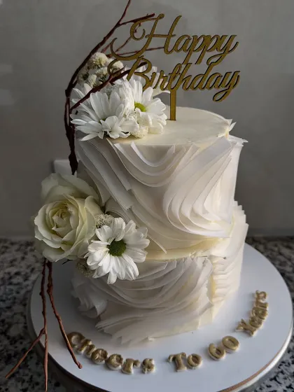 An elegant two-tier birthday cake featuring delicate white wafer paper ruffles and fresh white flowers. The design is complemented by rustic chocolate branches for a touch of nature.