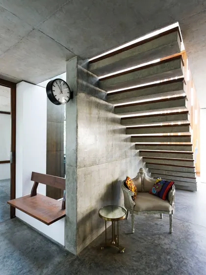 Light and material play a crucial role in our designs. Here, sunlight grazes a raw concrete wall, highlighting its texture, while a floating staircase with wooden treads adds warmth and lightness to the structure, creating a balanced and serene entryway.