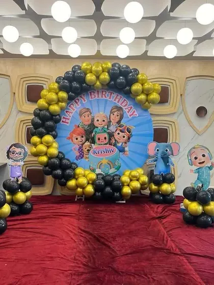 A Cocomelon themed birthday setup with a circular backdrop featuring the show's characters. The balloon arch is made of black and yellow balloons, with character props on balloon columns.