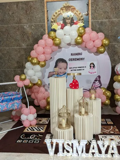 A complete view of a modern naming ceremony decor. This image shows the balloon arch, the personalized backdrop, the decorative pedestals, and the cradle, all coordinated in a pink and gold theme.
