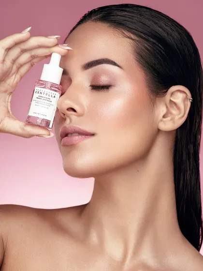 A serene and graceful shot featuring a model with eyes closed, holding a pore-minimizing serum. The image conveys a sense of calm and trust in the product. The soft pink background and gentle lighting enhance the healthy glow of her skin, creating a peaceful, spa-like mood.