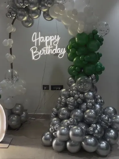 A modern and stylish birthday balloon arrangement in silver, white, and green. The abstract shape of the garland is paired with a simple 'Happy Birthday' neon sign.