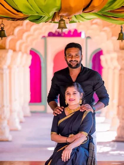 A happy couple poses on a decorated swing. Their classic black traditional outfits are beautifully highlighted by the colorful, festive backdrop.