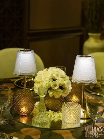 A complete tabletop centerpiece featuring a bouquet of white roses, elegant mini-lamps, and crystal candle holders on a mirrored base. This setup is the epitome of sophisticated dining.