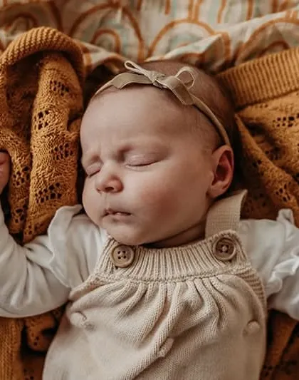 A close-up of a sleeping baby in a cute outfit, looking cozy under a warm blanket. We focus on comfort and safety during all our newborn sessions.